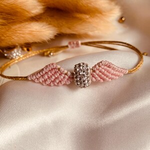 May include: A pink and gold friendship bracelet with a sparkly silver charm. The bracelet is made with a knotting technique and features a small, round charm with clear rhinestones.