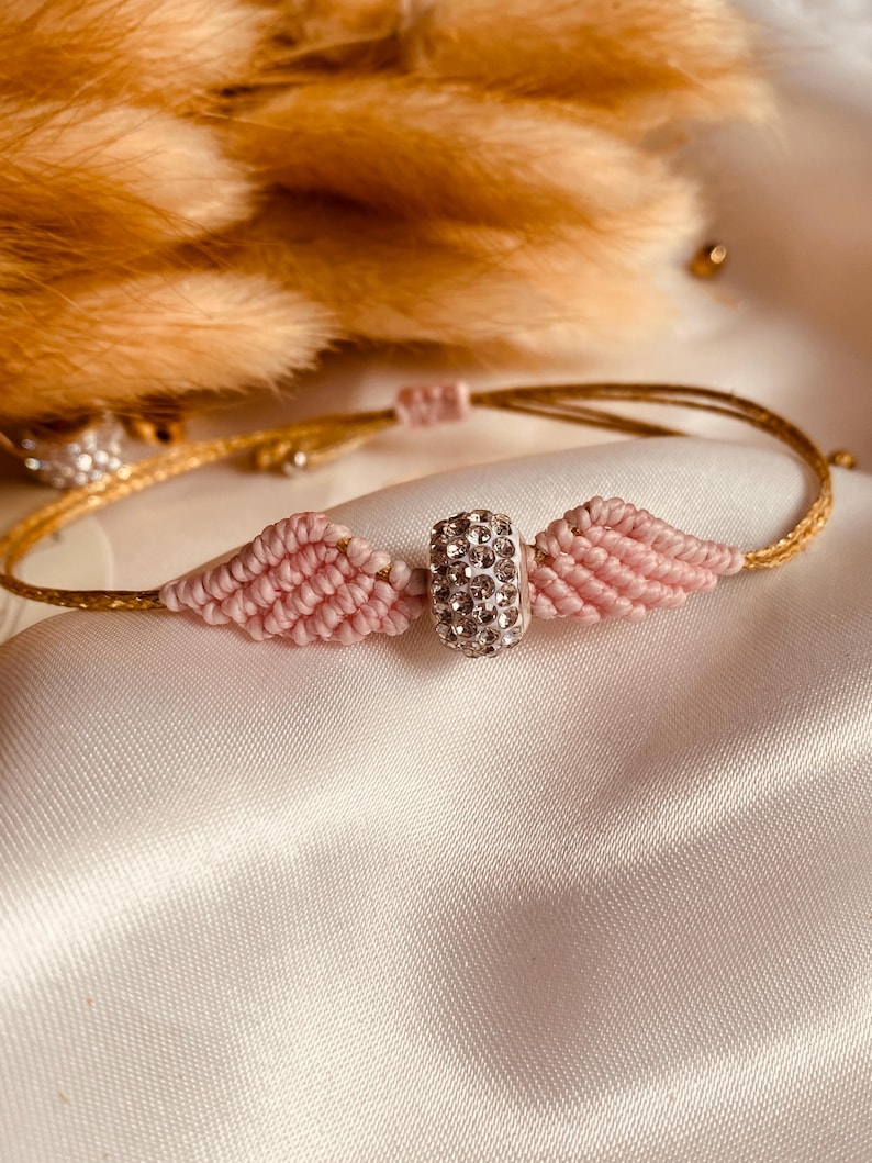 May include: A pink and gold friendship bracelet with a sparkly silver charm. The bracelet is made with a knotting technique and features a small, round charm with clear rhinestones.