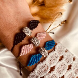 May include: Three adjustable friendship bracelets with a crystal charm in the shape of angel wings. The bracelets are black, pink, and blue.