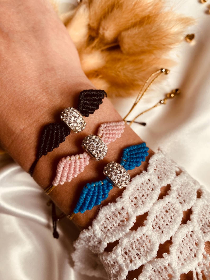 May include: Three adjustable friendship bracelets with a crystal charm in the shape of angel wings. The bracelets are black, pink, and blue.