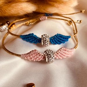 Pode incluir: Duas pulseiras de amizade com fio dourado e uma conta de cristal. Uma pulseira é rosa e a outra é azul.