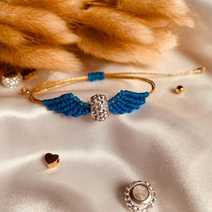May include: A blue and gold friendship bracelet with a crystal charm in the center. The bracelet features two blue wings on either side of the charm.