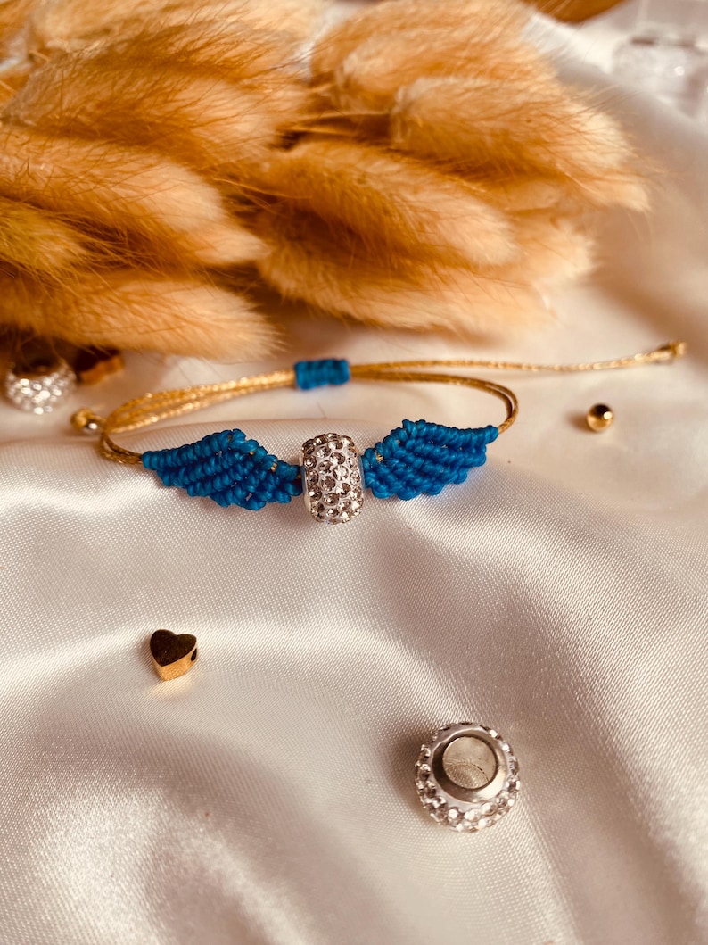 May include: A blue and gold friendship bracelet with a crystal charm in the center. The bracelet features two blue wings on either side of the charm.