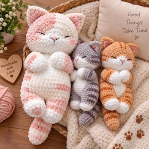 May include: Three plush crochet cats in pink and white, grey and white, and orange and white. Each cat has closed eyes, a small pink nose, and white paws. They are arranged together on a soft textured fabric inside a woven basket.
