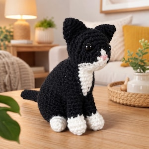 Könnte beinhalten: Eine handgehäkelte Katzenfigur aus schwarzem Garn für Körper und Schwanz, mit weißen Garnakzenten an Brust, Pfoten und Gesicht. Die Katze hat zwei große, runde, schwarze Kunststoffaugen und eine kleine, gestickte rosa Nase und sitzt aufrecht.