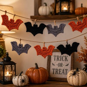 Peut inclure: Une guirlande décorative composée de sept chauves-souris crochetées en orange, noir et gris, suspendues à une fine ficelle. Les chauves-souris sont disposées sur deux rangées, mettant en valeur les textures du fil et les formes des ailes.