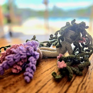 May include: Two crocheted figures, one with green hair and a green dress, the other with purple hair and a purple dress, lying on a wooden surface.