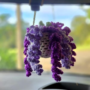 May include: A handcrafted crochet hanging basket in shades of purple and white. The basket is suspended by a chain and features cascading, textured crochet details. The design resembles a wisteria flower.