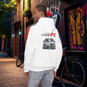 Könnte beinhalten: Weißer Kapuzenpullover mit dem Text "CIVIC TYPE R" und einer Autografik auf dem Rücken. Die Person trägt den Hoodie in einer städtischen Umgebung mit einer Backsteinmauer und Graffiti.