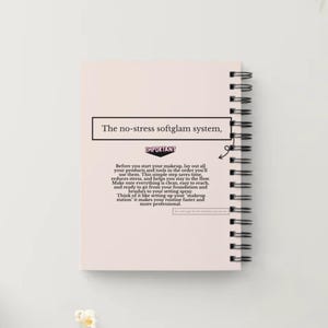 May include: A light pink spiral-bound notebook with the text "The no-stress softglam system," at the top. The notebook is open, revealing lined pages. A pencil and a bowl of popcorn are in the background. The notebook is on a white surface.