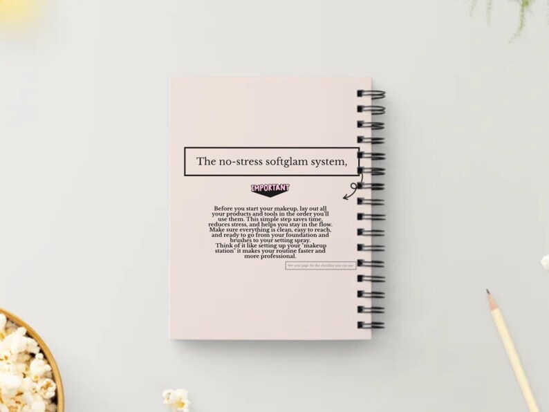 May include: A light pink spiral-bound notebook with the text "The no-stress softglam system," at the top. The notebook is open, revealing lined pages. A pencil and a bowl of popcorn are in the background. The notebook is on a white surface.
