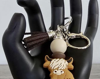 May include: A brown and white highland cow keychain with a brown tassel and silver beads. The keychain is attached to a silver key ring.