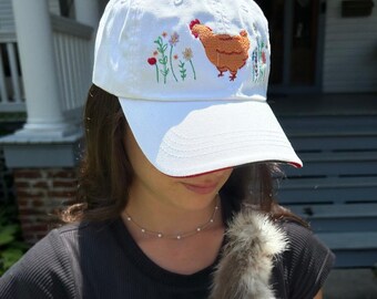 May include: White baseball cap with a red brim, embroidered with a brown chicken and colorful flowers. The cap is worn by a person holding a fluffy, gray and white Silkie chicken.