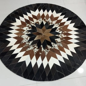 May include: A round rug with a geometric starburst design. The rug features a pattern of diamonds in black, white, brown, and tan cowhide. The center of the rug has a star shape with a mix of brown and tan cowhide.