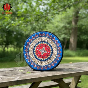 Puede incluir: Un objeto decorativo redondo, azul y negro, con un colorido diseño de mandala. El mandala presenta patrones rojos, amarillos y blancos. El objeto está colocado sobre una mesa de picnic de madera al aire libre.