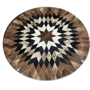 Peut inclure: Tapis rond avec un motif géométrique en forme d'éclat d'étoile. Le tapis présente un motif de losanges dans des tons de marron, noir et blanc. Le tapis est fait d'une matière naturelle, probablement du cuir, et a un aspect texturé.