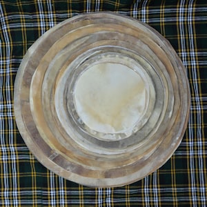 Op de afbeelding: Een stapel cirkelvormige trommelvellen van verschillende formaten, concentrisch gerangschikt. De vellen hebben een natuurlijke, lichtbruine kleur met donkere randen. De achtergrond is een groen, zwart en geel geruit patroon.