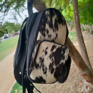 Puede incluir: Mochila de cuero negro con un bolsillo frontal y una sección superior de piel de vaca. La piel de vaca presenta un patrón de manchas negras sobre un fondo blanco. La mochila está colgada de una rama de árbol.