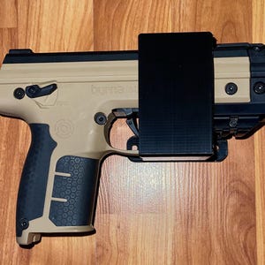 May include: Tan and black handgun with the text "byrna SD" visible on the side. The grip is black with a textured pattern. A black rectangular attachment is connected to the top of the handgun. The background is a wooden surface.