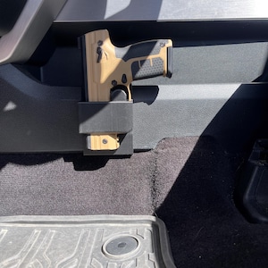 May include: A tan and black handgun is secured in a black holster mounted in the backseat of a vehicle. The holster is attached to the back of the seat.