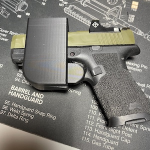 May include: A black handgun with an olive green slide and a black magazine holder. The handgun has a textured grip and a red dot sight. The image is on a black mat with text including "Barrel and Handguard".