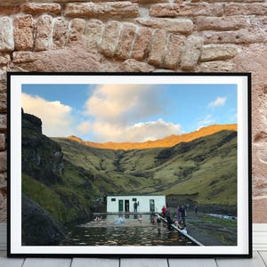 Puede incluir: Una fotografía enmarcada de una fuente termal natural en un valle con un pequeño edificio y personas nadando en el agua. El cielo es azul con nubes blancas y las montañas son verdes y marrones.