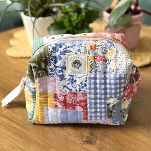 May include: A patchwork cosmetic bag with a gold zipper. The bag features a variety of floral and patterned fabrics in shades of blue, pink, yellow, and green. A white label with the text "Haven Rose Supply Co" is sewn on the front.