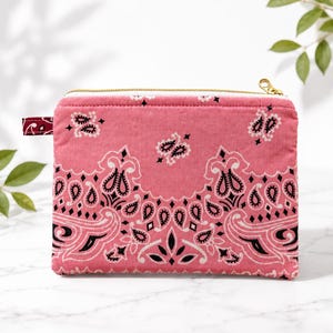May include: A rectangular zippered pouch made of pink fabric featuring a black and white paisley bandana print. The pouch includes a gold-toned metal zipper closure and a small matching fabric tab on the left side, showcasing a classic pattern.