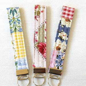 May include: Three fabric wrist keychains with gold-colored hardware. Each keychain features a unique patchwork design with floral, gingham, and geometric patterns in various colors, including blue, yellow, red, and white.
