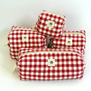 May include: A collection of red and white gingham patterned cosmetic bags in various sizes. Each bag features a gold zipper and a small, circular label with a floral design. The bags are arranged on a white background.