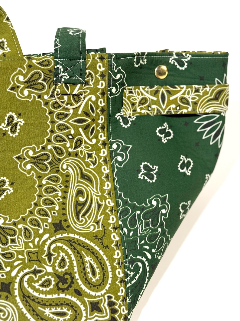 Puede incluir: Primer plano de un bolso tote con estampado de cachemira verde y verde oliva. El bolso presenta un dise&ntilde;o contrastante con un bolsillo y un cierre de bot&oacute;n a presi&oacute;n de metal. El bolso tiene un asa y un anillo de metal.