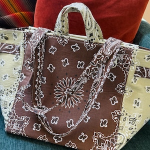 Puede incluir: Un bolso tote grande con un estampado de cachemira marr&oacute;n y blanco. El bolso presenta un panel central marr&oacute;n con un dise&ntilde;o floral blanco y paneles laterales de color marr&oacute;n claro. Las asas y las correas tambi&eacute;n tienen un estampado.