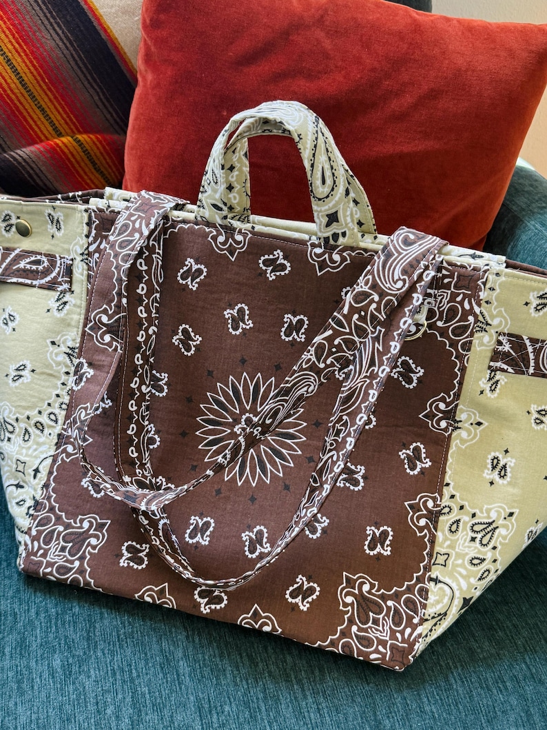 Puede incluir: Un bolso tote grande con un estampado de cachemira marr&oacute;n y blanco. El bolso presenta un panel central marr&oacute;n con un dise&ntilde;o floral blanco y paneles laterales de color marr&oacute;n claro. Las asas y las correas tambi&eacute;n tienen un estampado.