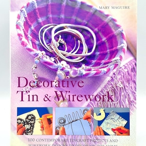 May include: A book cover titled "Decorative Tin & Wirework" by Mary Maguire. The cover features a purple basket filled with silver bracelets and jewelry. The book showcases 100 contemporary projects and designs for home decor.
