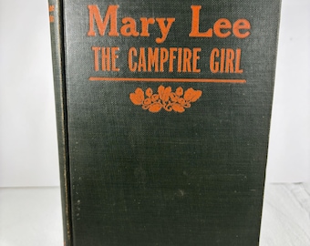 Op de afbeelding: Een vintage hardcover boek met de titel "Mary Lee The Campfire Girl". Het boek heeft een donkergroene kaft met oranje letters en decoratieve bloemelementen. Het boek wordt gepresenteerd op een transparante plastic standaard.