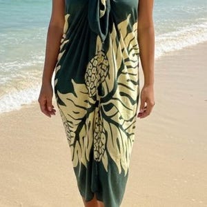May include: A dark green sarong with a yellow floral pattern, tied at the chest. The sarong is worn on a beach with the ocean in the background. The fabric drapes to the ankles.