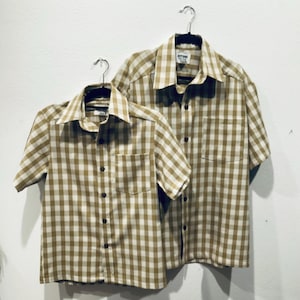 May include: Two gingham shirts in a brown and white pattern, hanging on hangers. The shirts have short sleeves and button-up fronts.