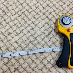 May include: A close-up of a beige woven fabric with a yellow rotary cutter and a white measuring tape. The tape measures up to 10 inches.
