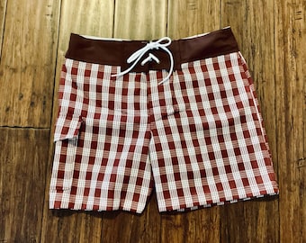 May include: Red and white plaid swim trunks with a brown waistband and white drawstring. The trunks have a side pocket.