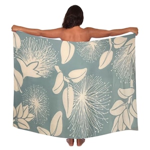 May include: A blue-green beach towel with a floral pattern. The design features large, cream-colored flowers and leaves. The towel is held up, showcasing the full pattern and color scheme.