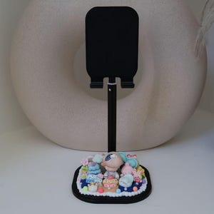 May include: A black phone stand with a decorative base featuring a colorful cake with cartoon characters. The stand is made of metal and has a simple design.