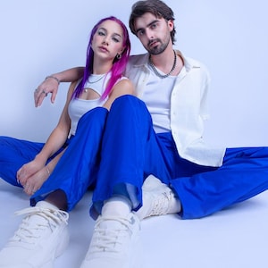 May include: Two people are sitting on a white background. The person on the left has pink and purple hair and is wearing a white tank top, blue pants, and white sneakers. The person on the right has brown hair and is wearing a white shirt, blue pants, and white sneakers.