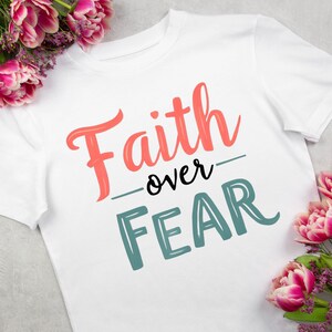 May include: White t-shirt with the words "Faith over Fear" in coral and teal lettering. The word "Faith" is in a script font, and "Fear" is in a block font. Pink and purple flowers are in the background.