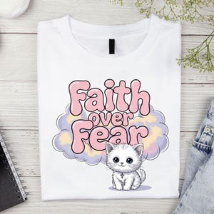 Könnte beinhalten: Weißes T-Shirt mit einem rosa und violetten Wolken-Design und einem weißen Kätzchen mit einem Kreuz-Halsband. Der Text "Faith over Fear" ist in rosa Buchstaben geschrieben.