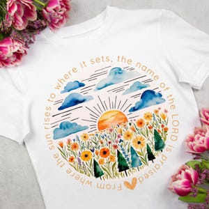 Puede incluir: Camiseta blanca con una ilustración de acuarela de una salida del sol sobre un campo de flores. El texto "From where the sun rises to where it sets, the name of the LORD is praised" está impreso en un círculo alrededor de la ilustración.