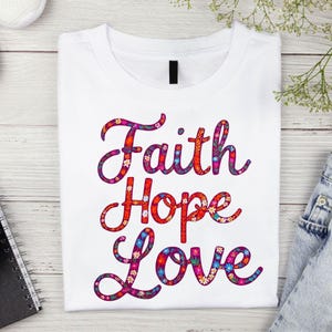 Op de afbeelding: Wit T-shirt met de woorden "Faith Hope Love" in een bloemenontwerp. De woorden zijn in een cursief lettertype geschreven en zijn paars omrand. De bloemen zijn in tinten roze, rood, oranje en geel.