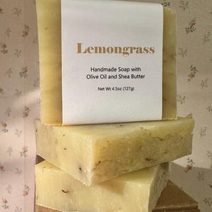 May include: Three bars of handmade lemongrass soap are stacked on top of each other. The top bar has a white label with the word "Lemongrass" in gold lettering. The label also states the soap is made with olive oil and shea butter, and weighs 4.5oz (127g).