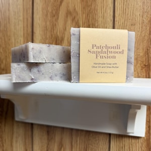 Peut inclure: Deux pains de savon rectangulaires gris clair faits à la main avec des taches sombres visibles. Un pain est enveloppé dans une étiquette en papier beige indiquant Patchouli Sandalwood Fusion, Handmade Soap with Olive Oil and Shea Butter, Net Wt 4.5oz (127g).