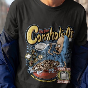 May include: A black t-shirt with a cartoon illustration of a character from the animated series "The Great Cornholio's" holding a spoon over a bowl of cereal. The text "The Great Cornholio's" is in yellow and the text "Are you threatening me?" is in white. The text "Free TP Inside!" is in a red and yellow speech bubble.
