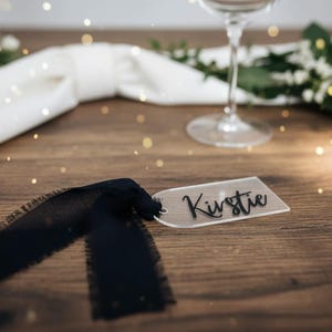 May include: A clear acrylic name tag, inscribed with "Kirstie" in black script, is paired with a black ribbon with a frayed edge. The tag sits on a wooden surface, with a wine glass and floral decorations in the background.